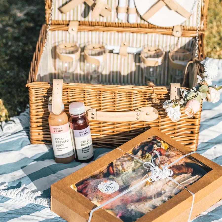 Picnic basket, coloured juices and assorted foods in a box on top of picnic blanket
