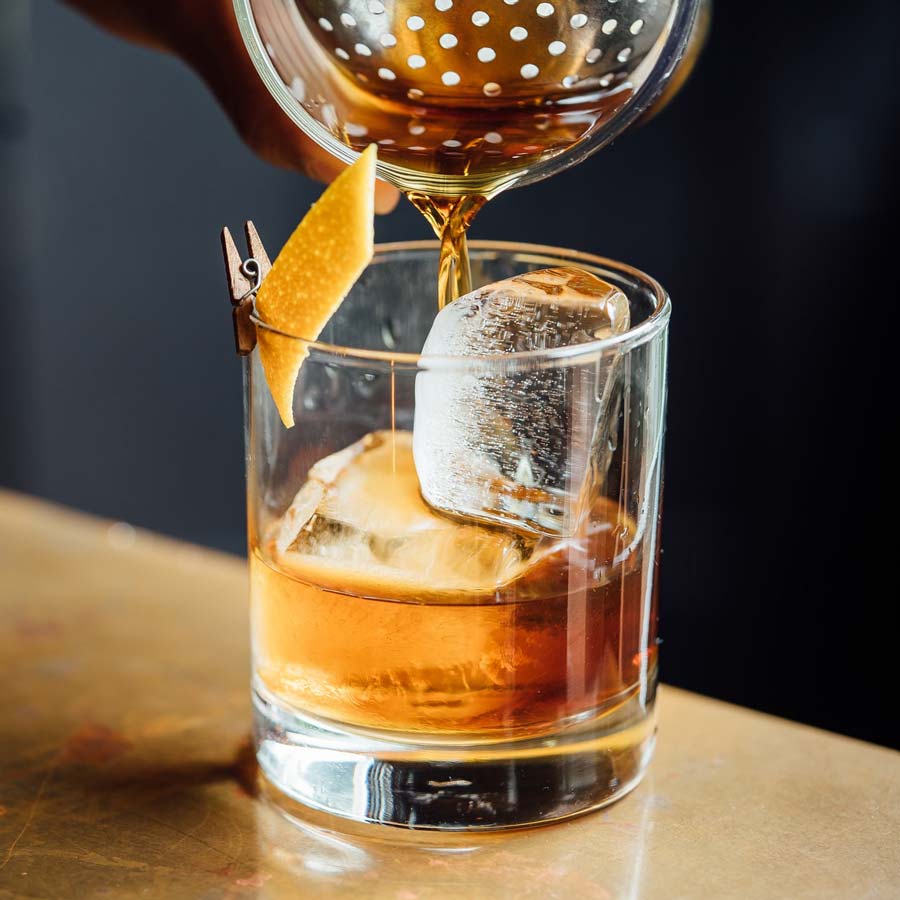 Pouring old fashioned into glass with ice and orange peel
